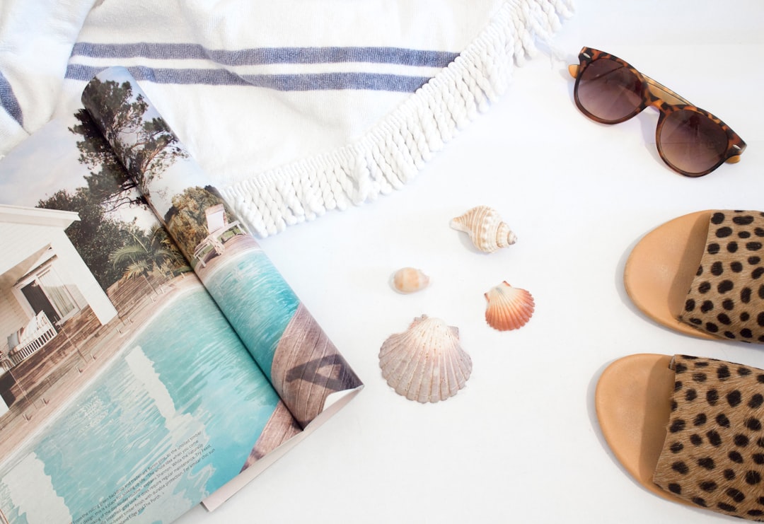 opened magazine beside seashells, slide sandals, sunglasses and towel on white surface